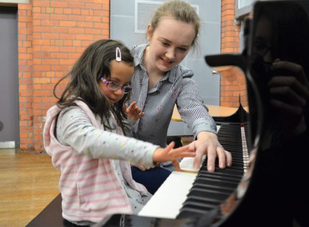 A girl wearing glasses tries to play notes on the piano with the help of an adult